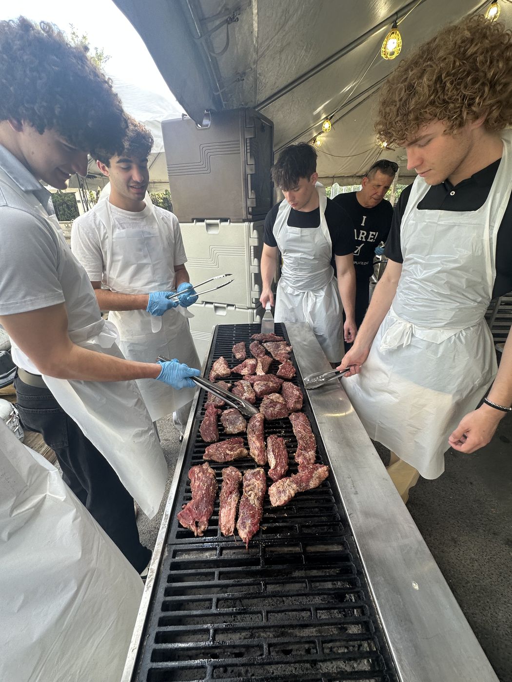 Steaks searing on a long grill as volunteers work them with tongs