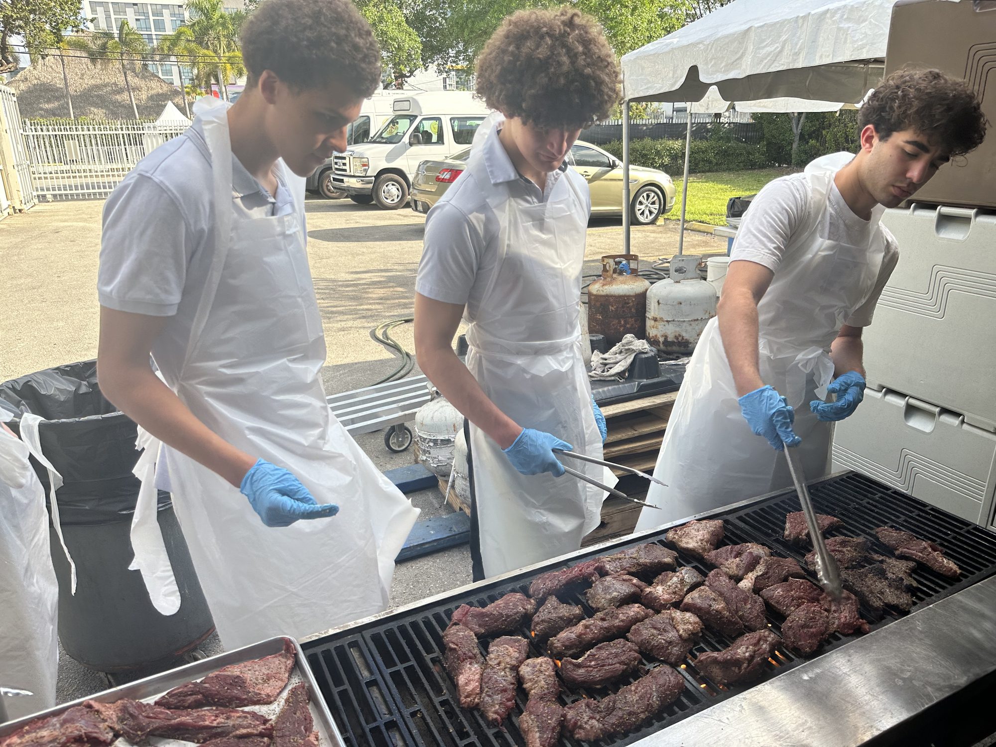 Three Giving Grill volunteers in aprons cooking steaks on an outdoor grill at a Miami Steak Night